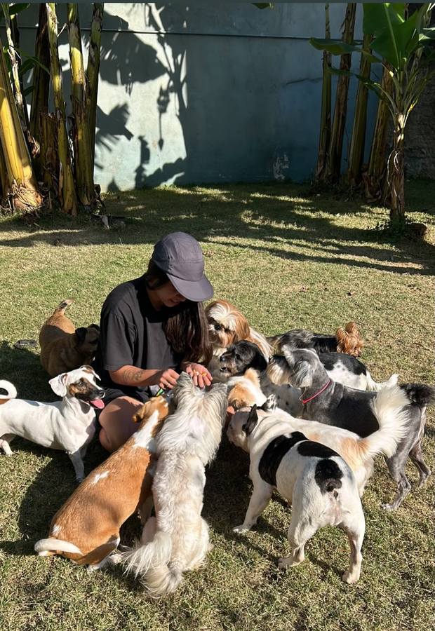 Uma das tutoras sentada na grama, rodeada por cerca de dez cães da creche